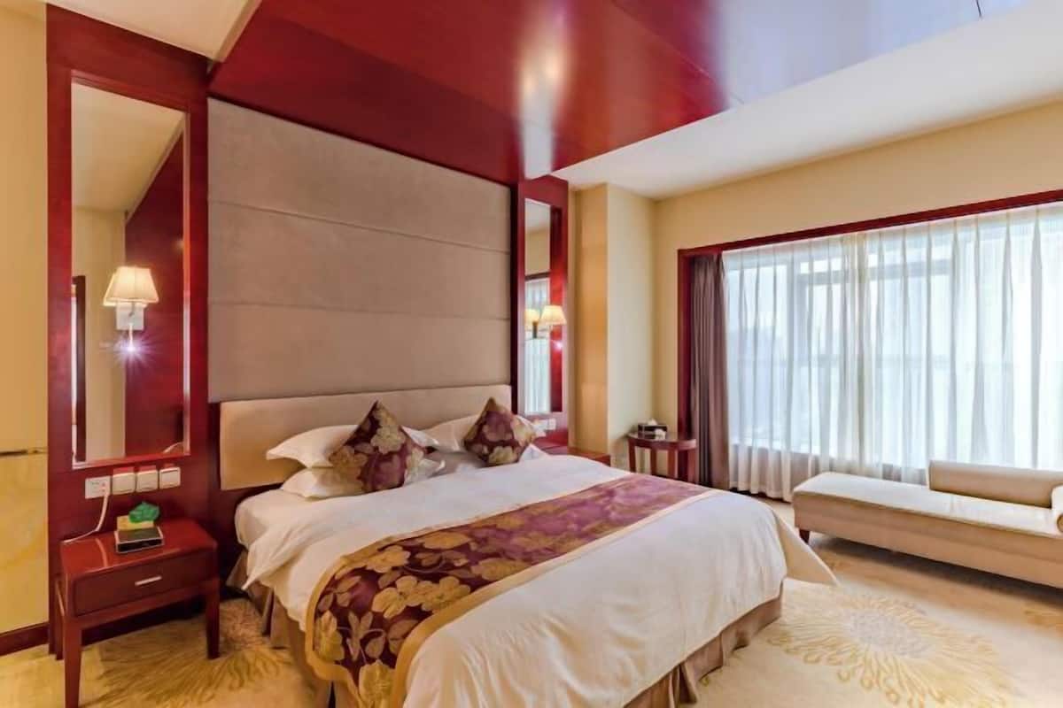 A hotel room with a large bed, a bedside table with a lamp, a sofa, and a large window with curtains.