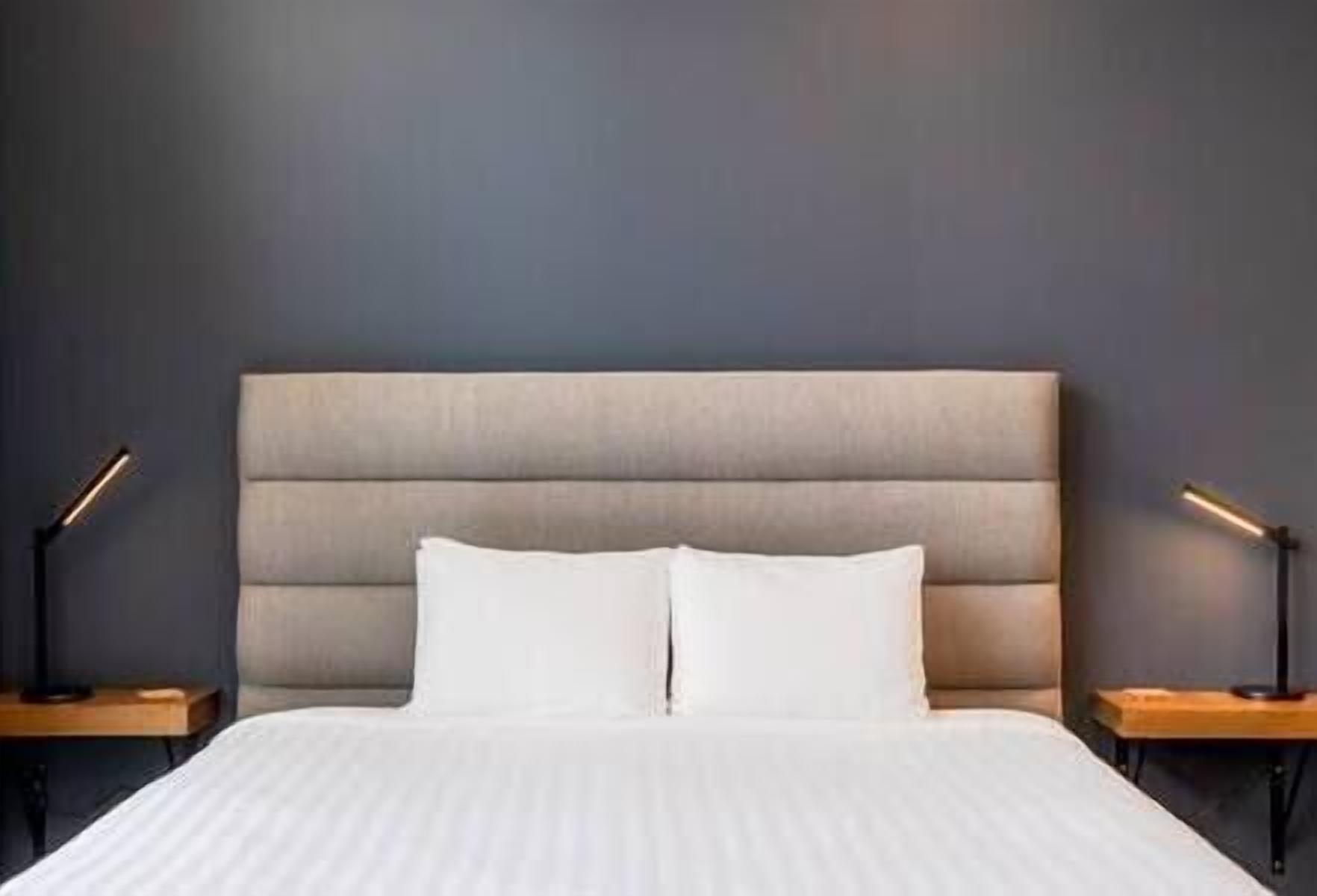 Een netjes opgemaakt bed met een grijs gestoffeerd hoofdbord, twee nachtkastjes met lampen en een effen grijze muur.