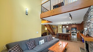 A spacious living area with a gray sofa, wooden coffee table, and a stone accent wall. A kitchen with modern appliances and a stone fireplace is visible in the background. - 3 BR Luxury Townhouse / Mountain views (Fernie)