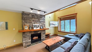 A living room with a stone fireplace, a mounted TV, a large window, and a sofa. - 3 BR Luxury Townhouse / Mountain views (Fernie)