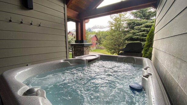 A hot tub with a stone surround, wooden ceiling, and a view of a house and trees. - 3 BR Luxury Townhouse / Mountain views (Fernie)