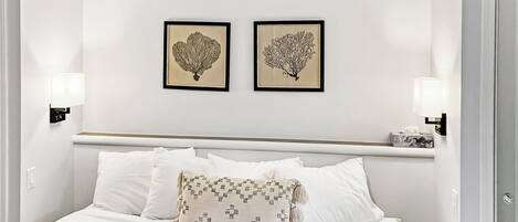 Una cama bien tendida con sábanas blancas y una almohada decorativa, cabecera con acabado en madera clara, y dos láminas botánicas enmarcadas en la pared.