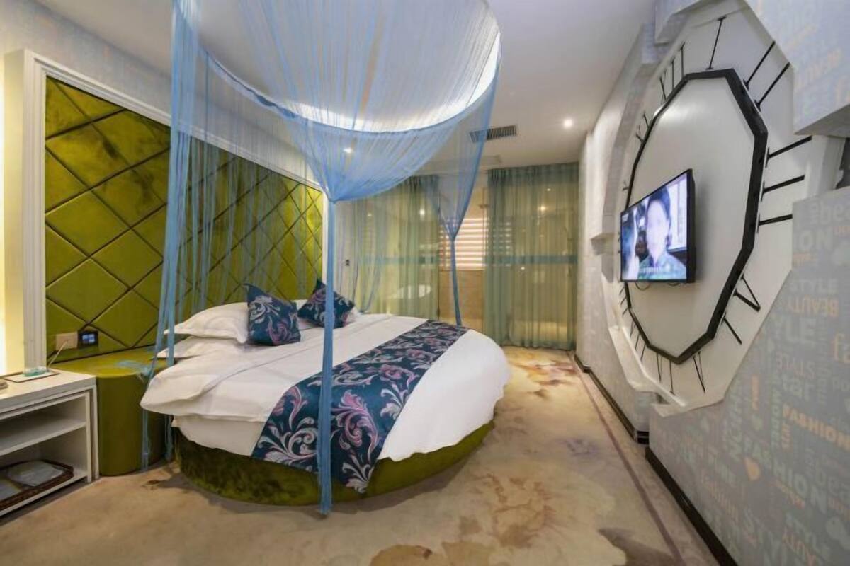 Une chambre moderne avec un lit placé sous un dais bleu, une grande horloge murale et un téléviseur fixé au mur.