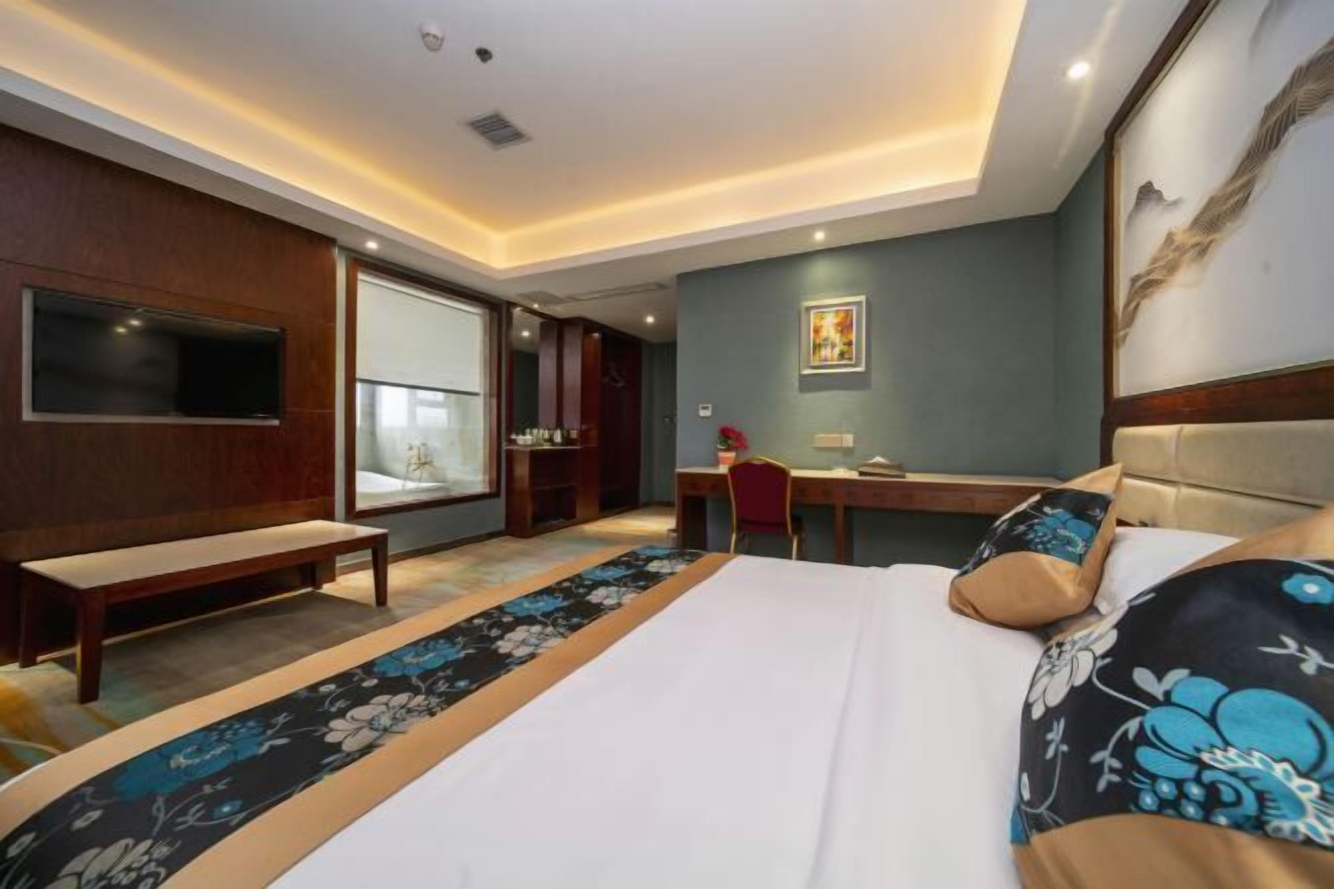 Een hotelkamer met een bed, een bureau, een stoel, een bankje, een televisie en een groot raam.