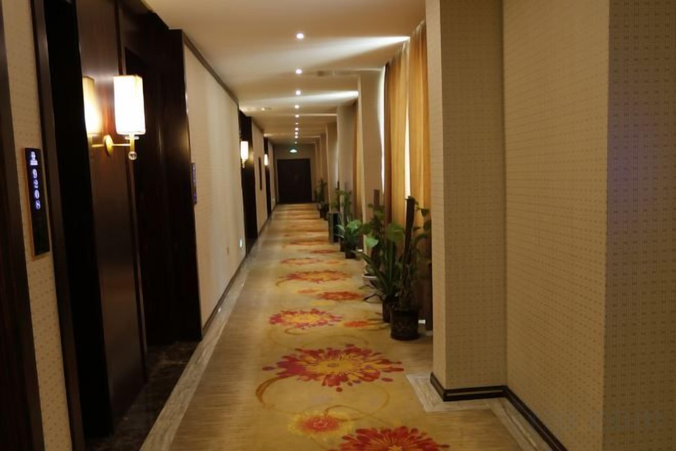 A hotel corridor with a carpeted floor, wall-mounted lamps, and potted plants.