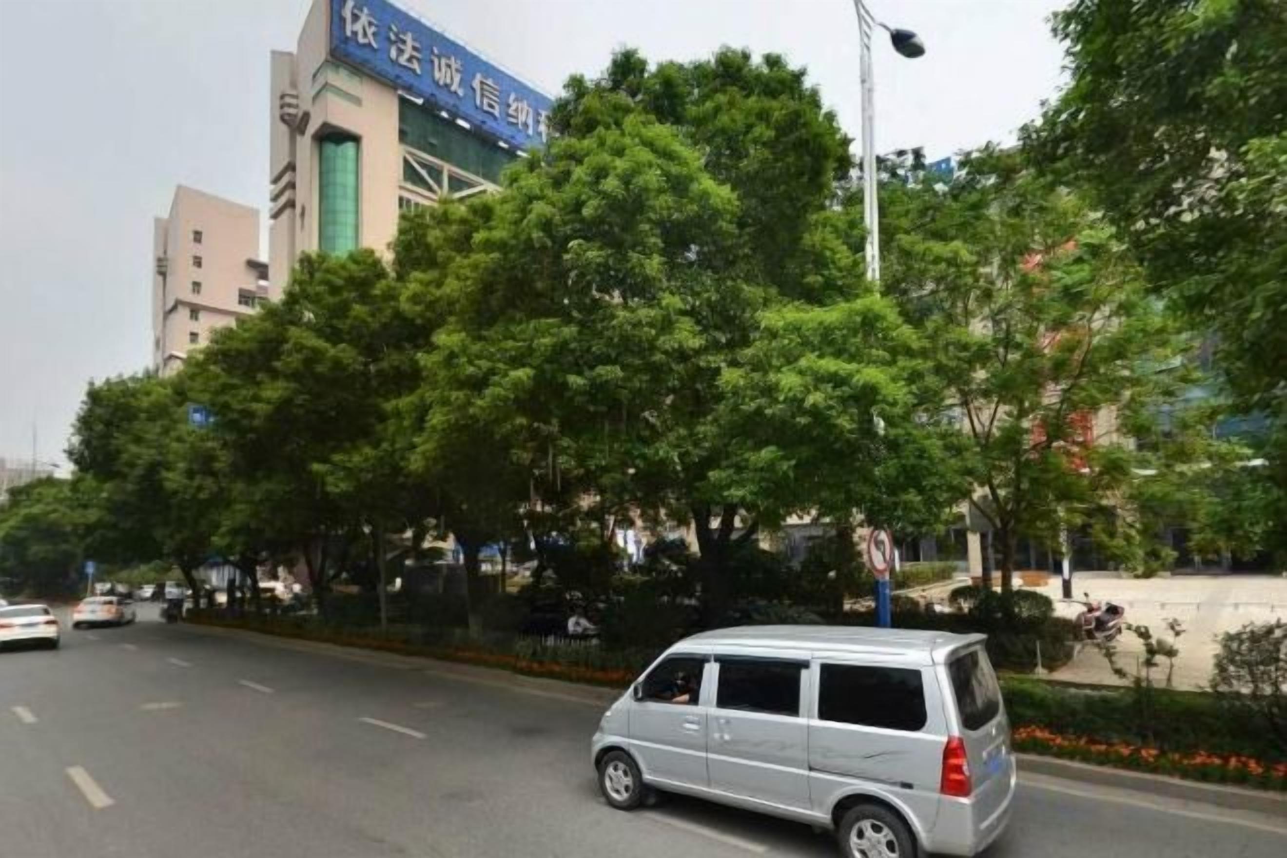 A street scene with a silver minivan parked on the side of the road, trees lining the street, and a building with a blue sign in the background.