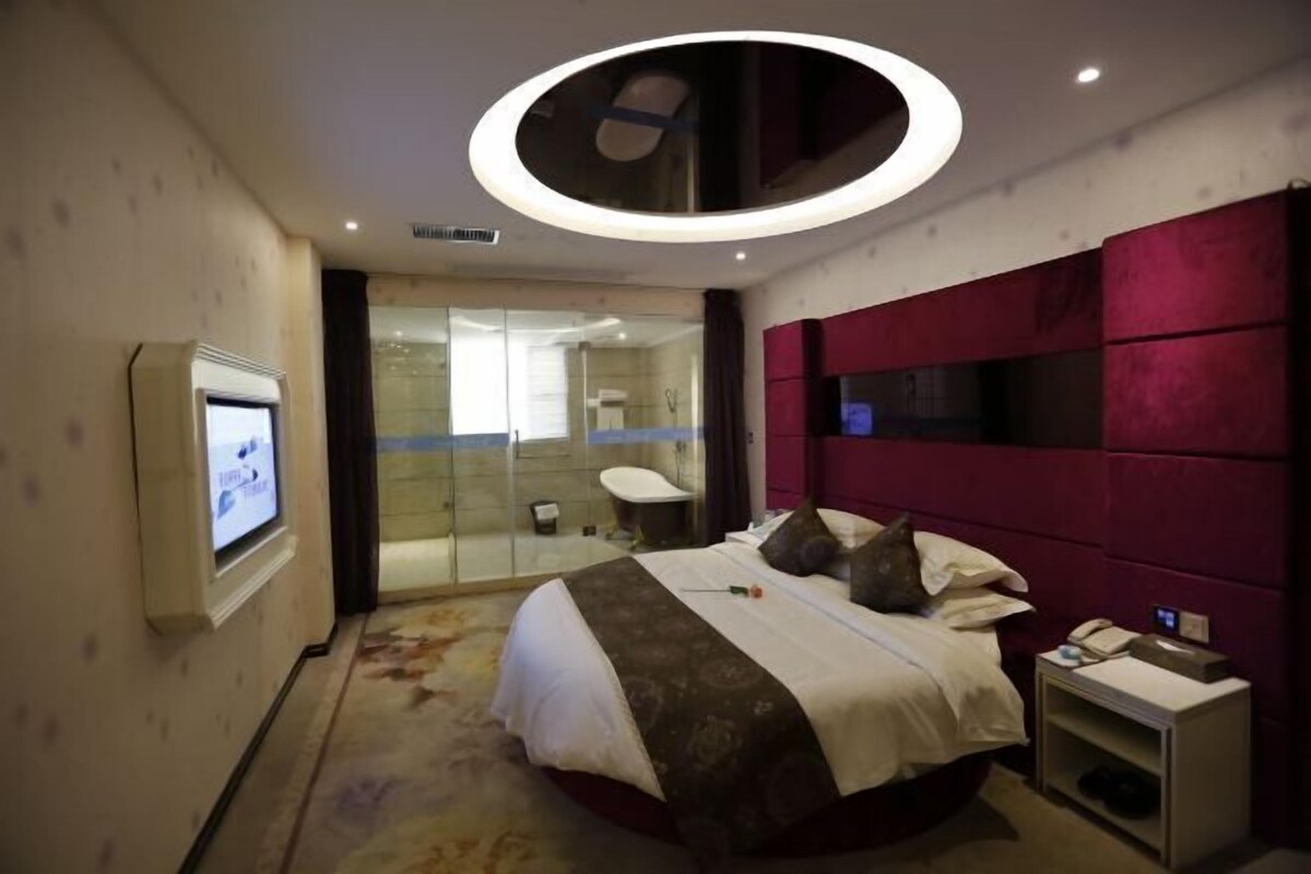 A modern hotel room with a large bed, a TV, a glass shower enclosure, and a red accent wall.
