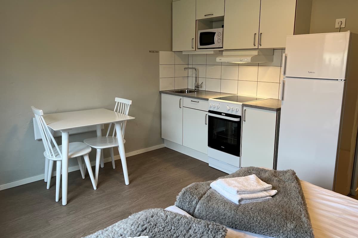 Een compacte keuken met witte kastjes, een magnetron, oven en een koelkast. Er is een kleine eettafel met stoelen.