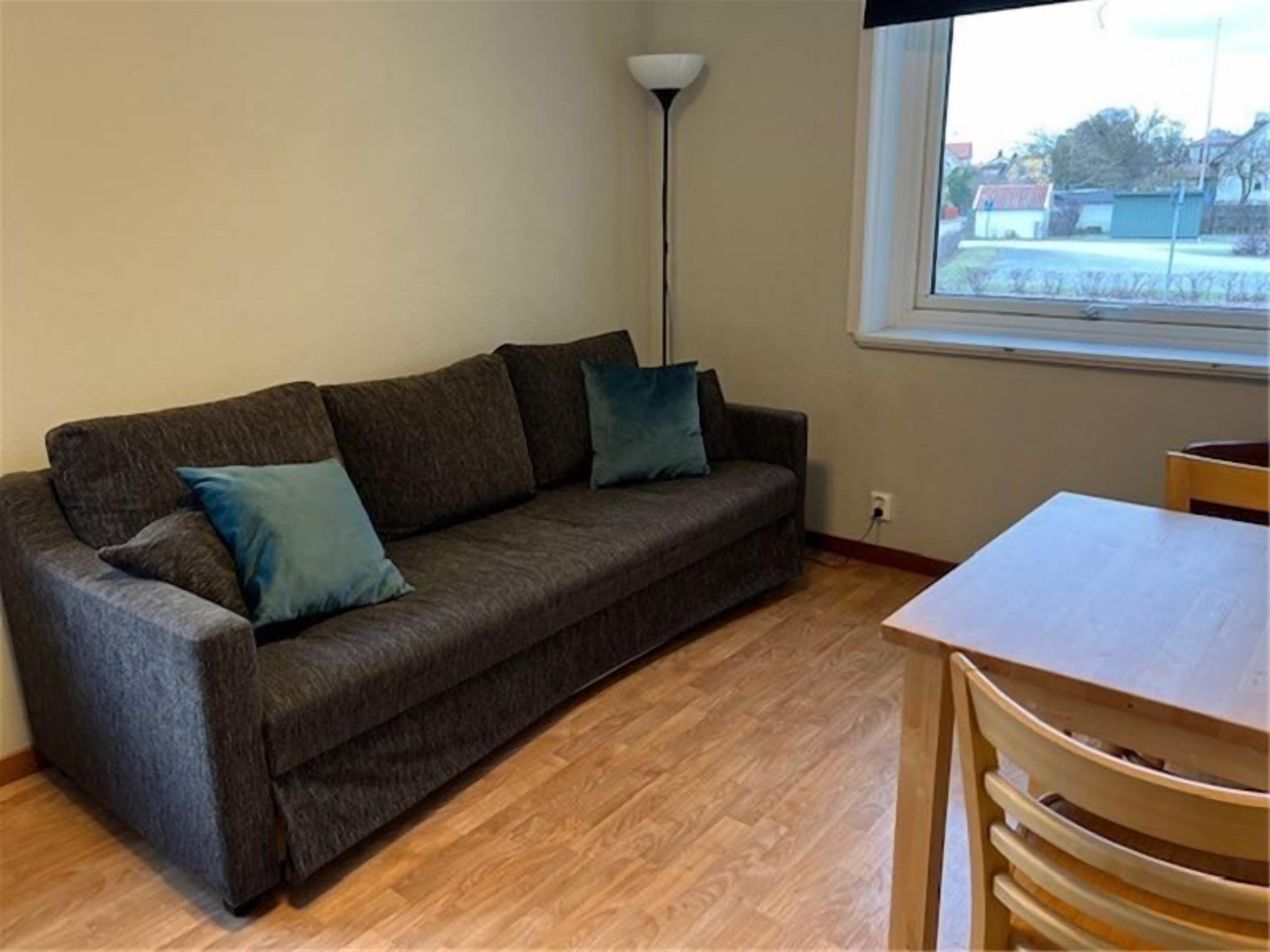 Una sala de estar con un sofá, dos almohadas, una lámpara de pie, una mesa y un ventanal con vistas a un edificio.