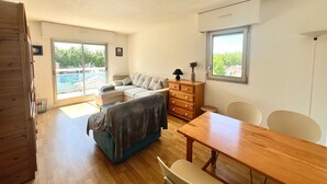 A spacious living area with a sofa, a wooden dresser, a dining table, and a view of the outdoors through a large window. - "Charming Apt near Casino & Beach, Parking, Balcony, Shops Nearby" (Arcachon)