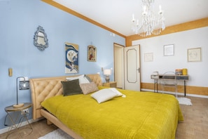 A bedroom with a bed, a chandelier, a desk, and artwork on the walls.
