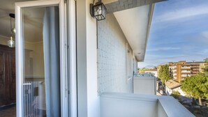 En balkon med glasdør, flisebeklædt væg og udsigt til bygninger og træer.