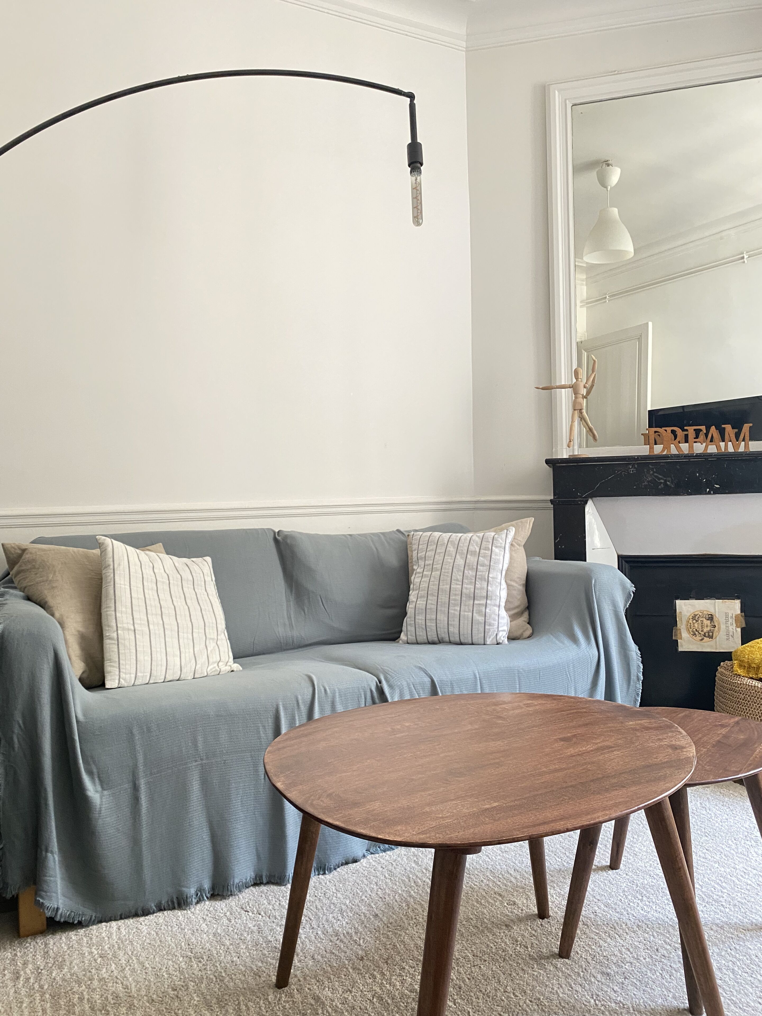 Un salon avec un canapé gris, deux tables basses en bois, un lampadaire noir et une cheminée surmontée d’un miroir.