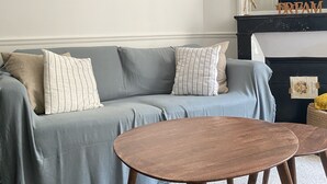Een woonkamer met een grijze bank, twee houten salontafels, een zwarte vloerlamp en een open haard met een spiegel erboven.