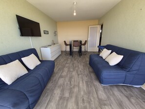 A room with two blue sofas, a white dresser, a TV mounted on the wall, a dining table with chairs, and a door leading to another room.