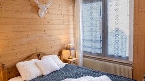 Ein Schlafzimmer einer Holzhütte mit einem Bett, einem Nachttisch, einer Lampe, einer Dekoration mit Hirschkopf und einem Fenster mit herzkugeligem Vorhang.