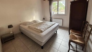 Een slaapkamer met een houten plafond, een bed met quilt, een raam met gordijnen, een radiator en een stoel.