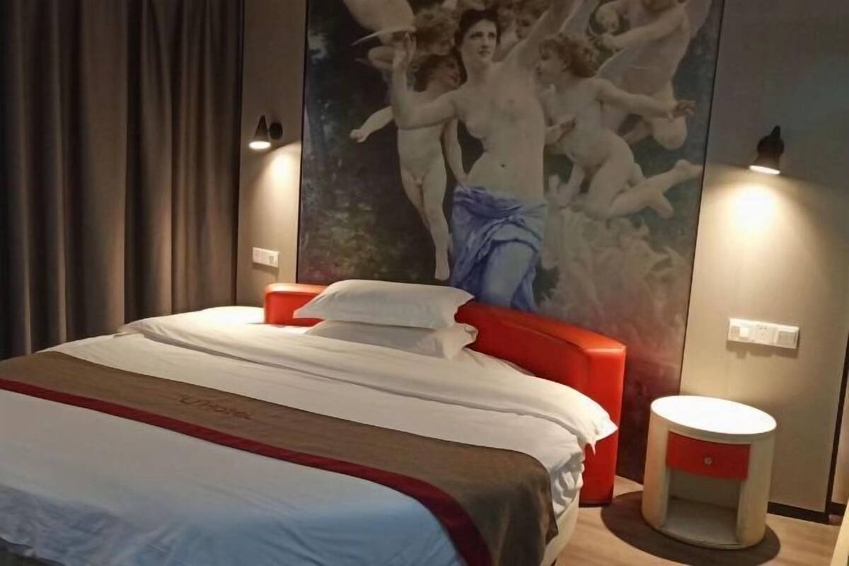 A hotel room with a large bed, a red accent chair, a round bedside table, and a wall with a large painting of cherubs.