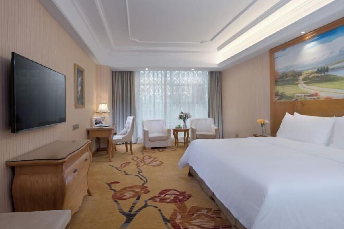 A hotel room with a large bed, a desk with a chair, a television, and a painting on the wall.