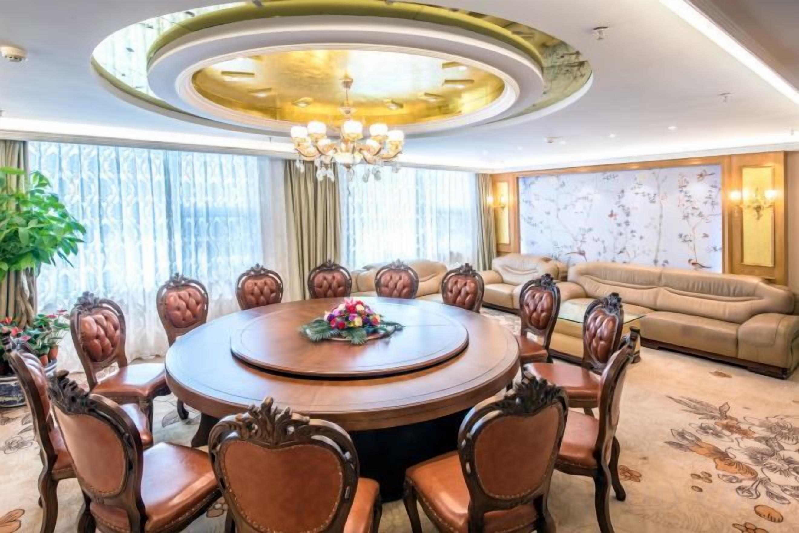 A round dining table with chairs, a chandelier, and a sofa in a well-lit room with large windows.