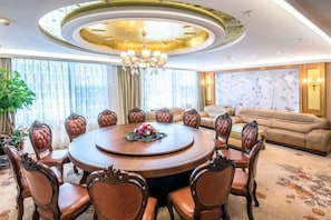 A round dining table with chairs, a chandelier, and a sofa in a well-lit room with large windows.
