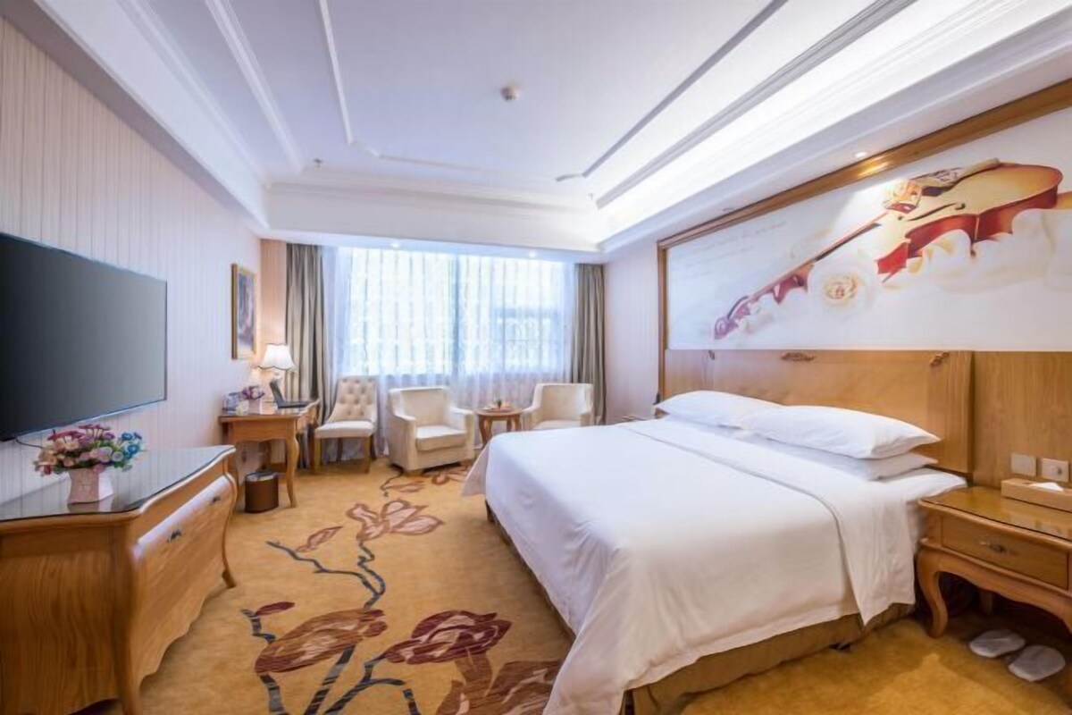 A hotel room with a large bed, a flat-screen TV, a desk with a lamp, a chair, and a window with curtains.