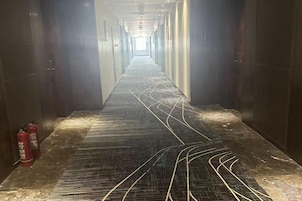 Un pasillo largo y estrecho con piso estampado, extintores a la izquierda y puertas a ambos lados.