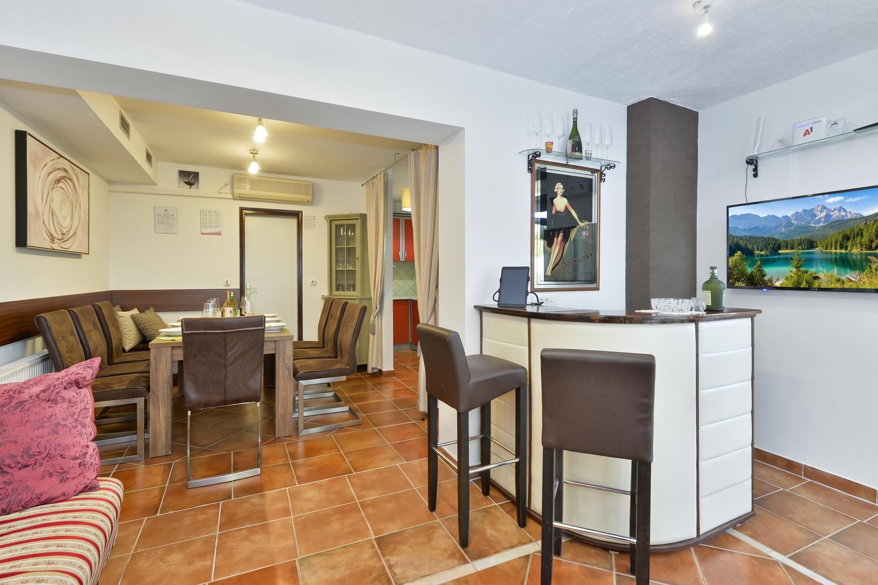 Una sala de estar moderna con comedor, barra y televisor mostrando un paisaje montañoso.