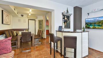 Una sala de estar moderna con comedor, barra y televisor mostrando un paisaje montañoso.