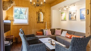 A cozy room with a wooden ceiling, a chandelier, a sofa with cushions, wicker chairs, a table with a remote control, and artwork on the walls. - Chalet # 22 with 4 bedrooms & sauna (Murau)
