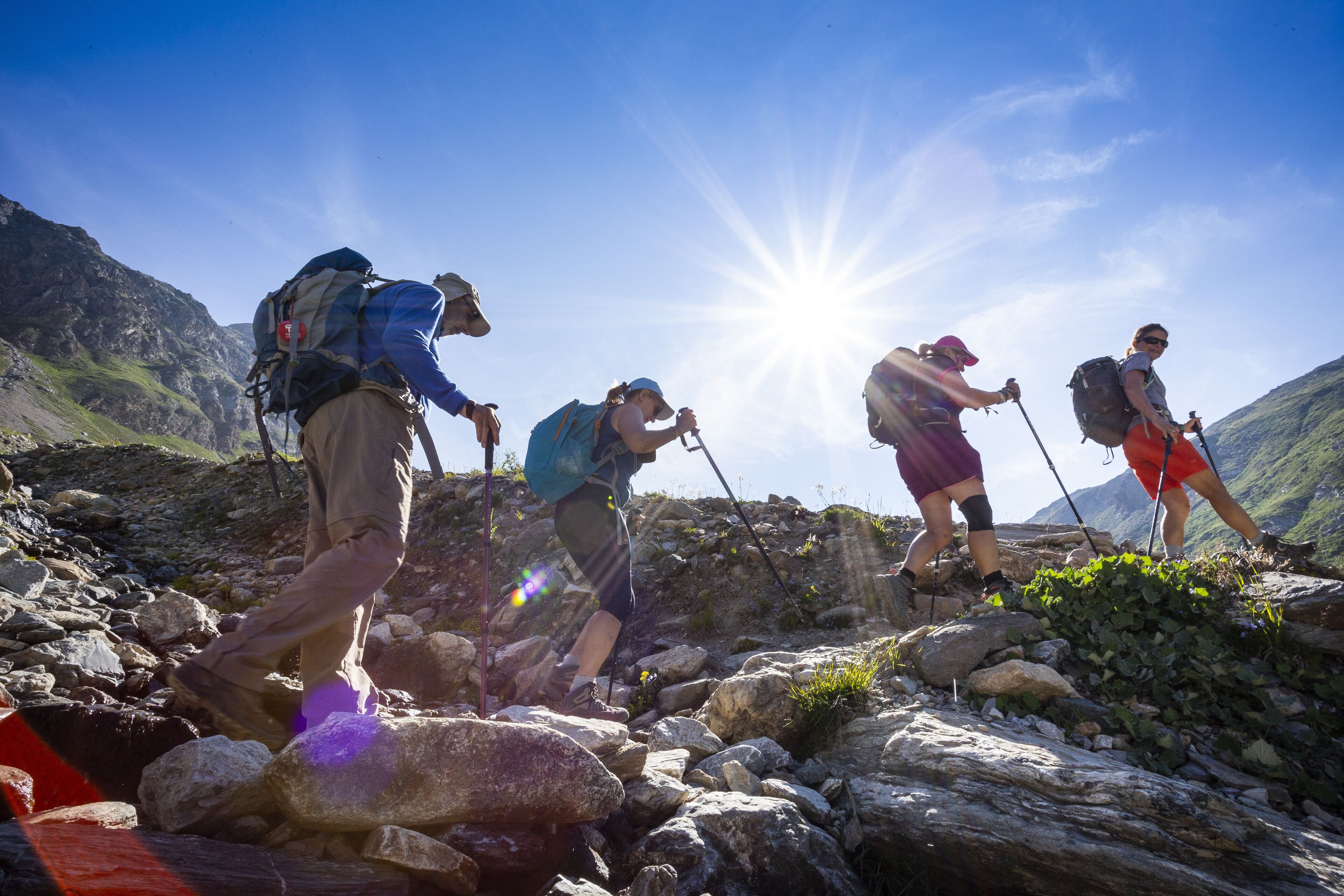 Un groupe de randonneurs sur un sentier de montagne rocailleux, sous un ciel dégagé et un soleil éclatant.