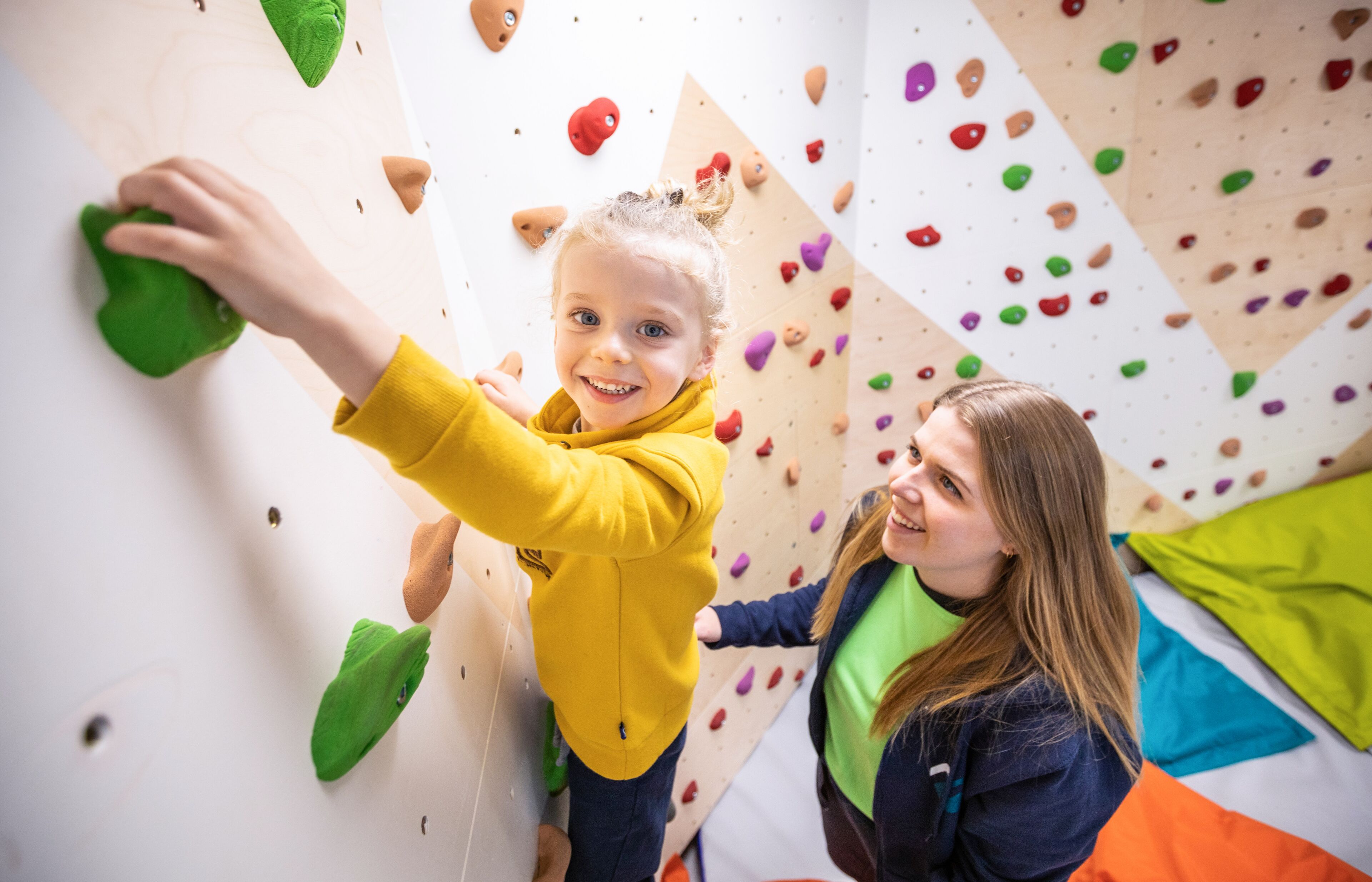 A child climbing a wall with colorful holds, and an adult standing beside them.