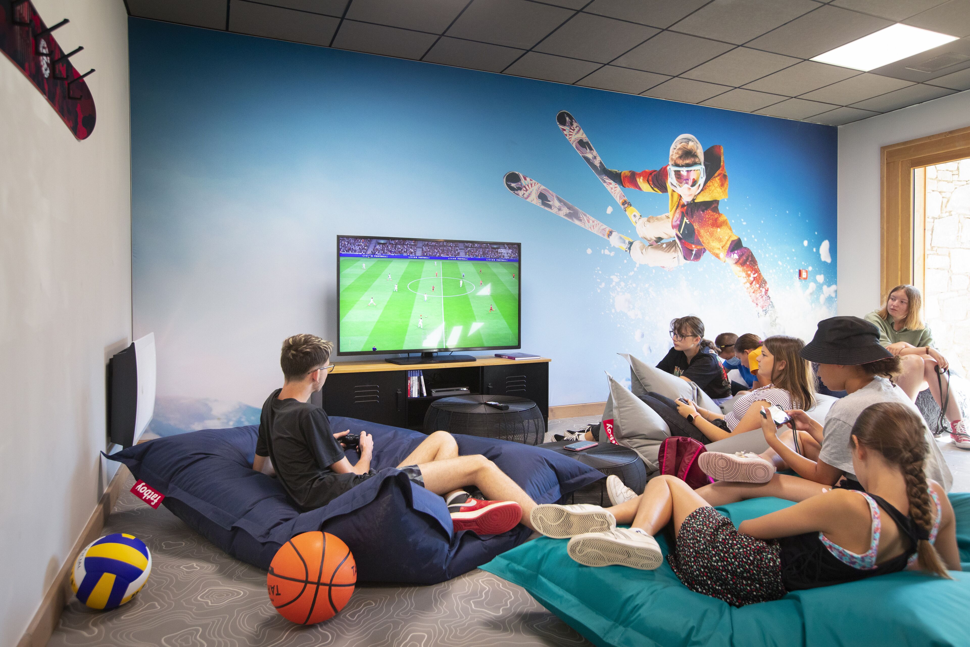 Eine Gruppe von Menschen schaut ein Sportspiel auf einem großen Fernseher in einem Raum mit einem blauen Wandgemälde und einem Fenster.
