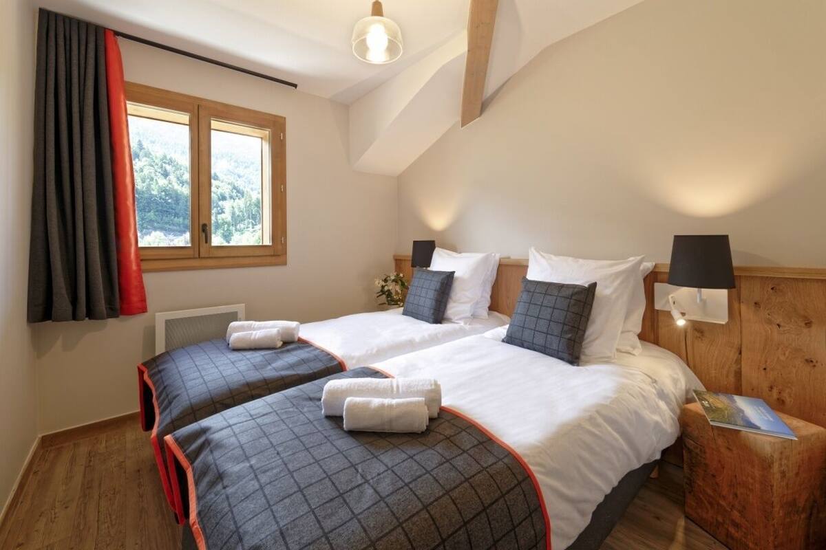 Une chambre avec deux lits, une fenêtre avec des rideaux, une lampe de chevet et une tête de lit en bois.