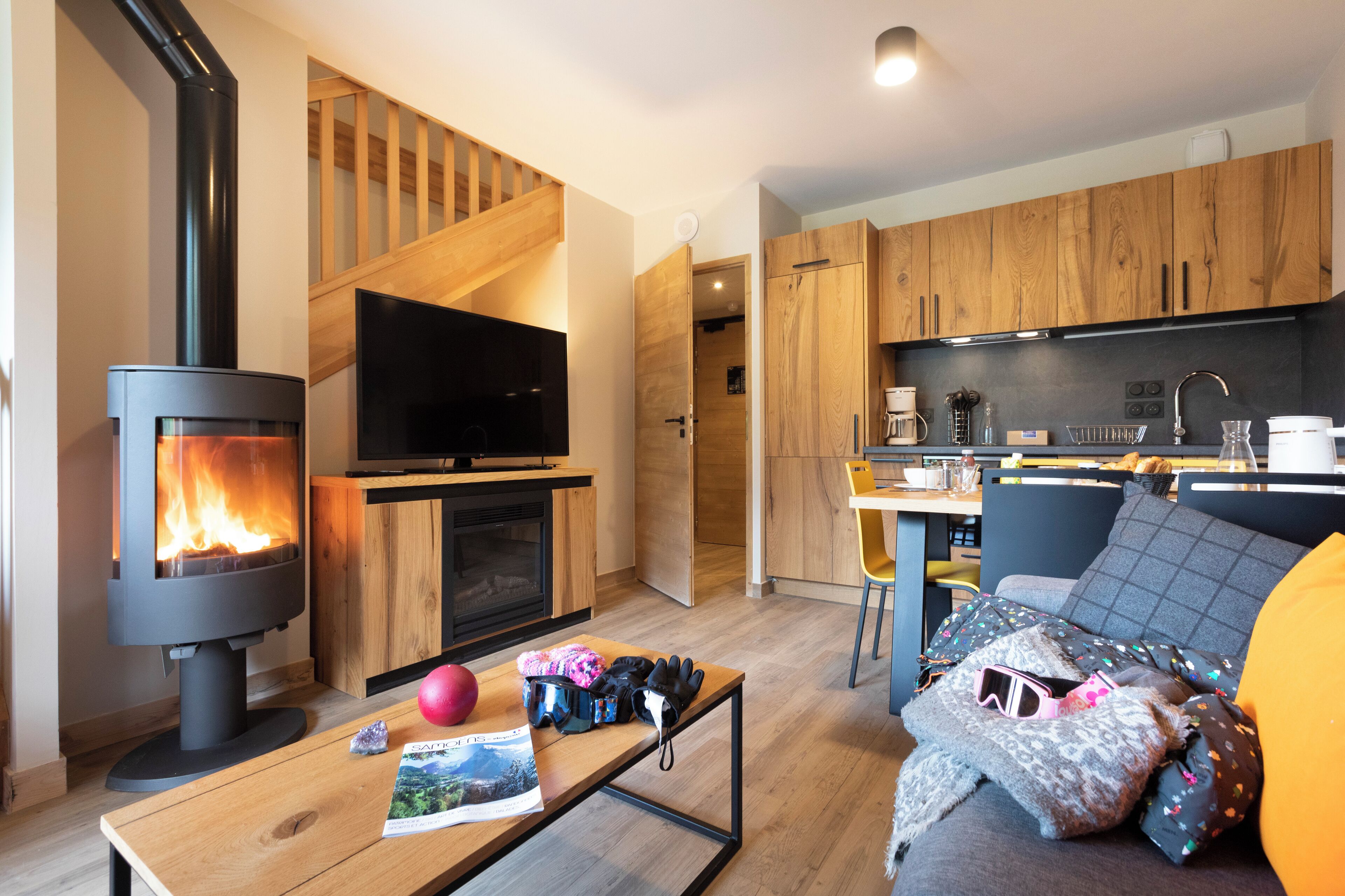 A modern living room with a wood-burning stove, a flat-screen TV, a dining area with a bar, and a kitchen with wooden cabinets.