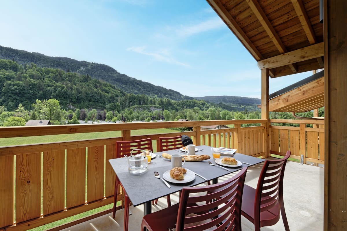 Une terrasse en bois avec une table dressée pour le petit-déjeuner, offrant une vue sur un paysage montagneux.