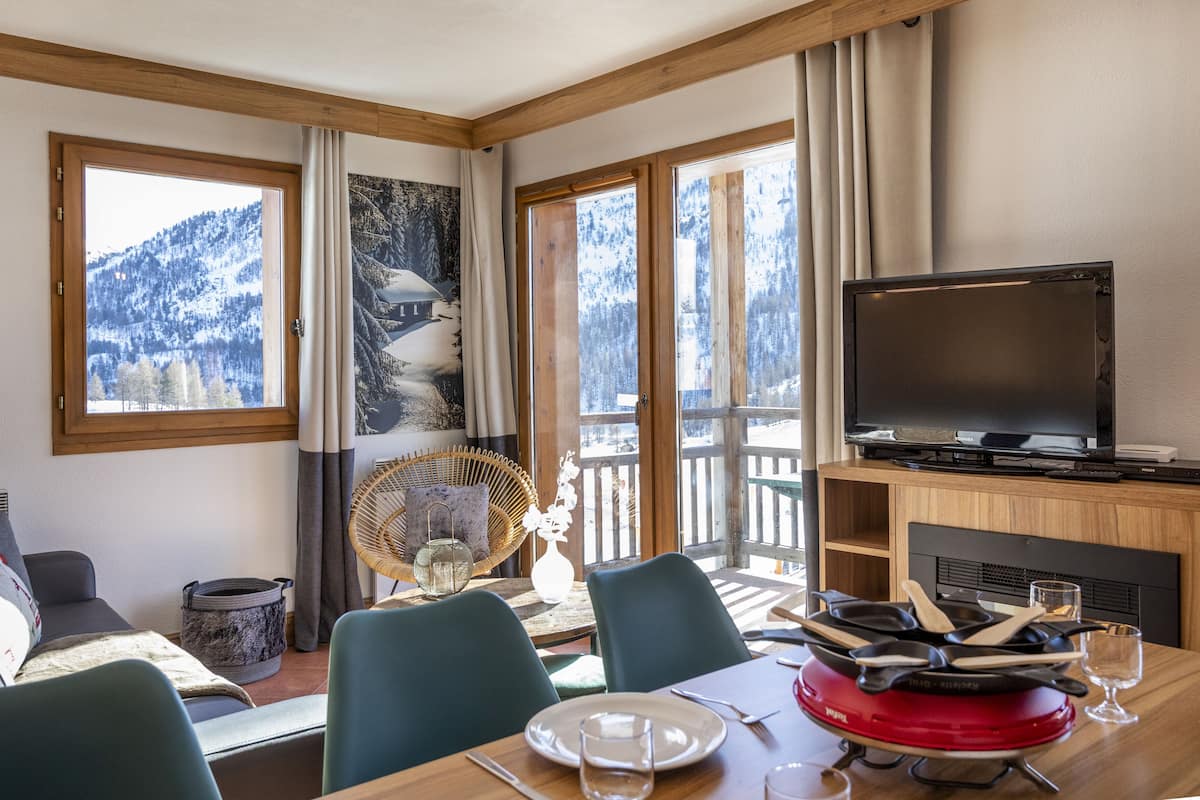 Un coin repas avec une table dressée pour deux personnes, un téléviseur et une vue sur des montagnes enneigées, visible à travers de grandes fenêtres.
