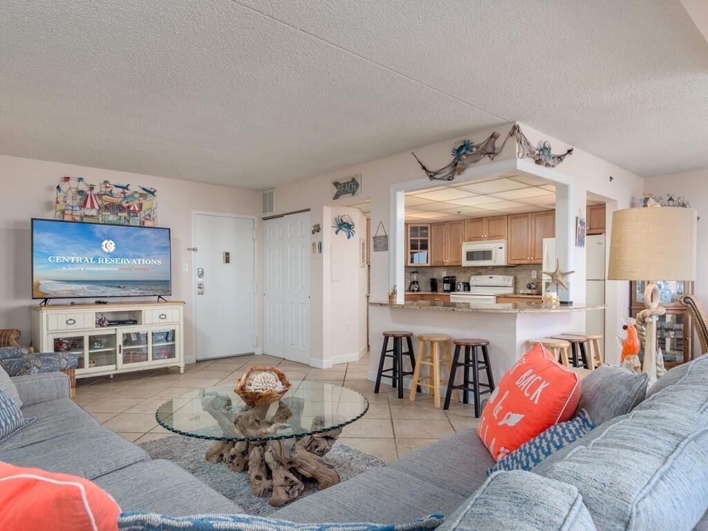 A living room with a television, sofa, and coffee table. There is a kitchen area visible through an open door.