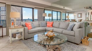 A modern living room with a grey sectional sofa, a glass coffee table, and a view of the city through large windows.