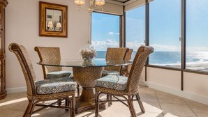 A dining area with a round glass table surrounded by wicker chairs, large windows offering a view of the ocean, and a chandelier hanging from the ceiling.