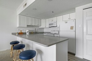 A modern kitchen with white cabinets, a grey countertop, a microwave, a refrigerator, and three blue bar stools.