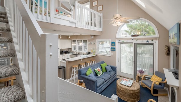 A spacious loft apartment with a mezzanine level, a kitchen, a living area with a sofa and chairs, and a large window providing natural light.