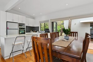 Een moderne keuken met witte kasten, een eettafel met houten stoelen en uitzicht op de omgeving door grote ramen.