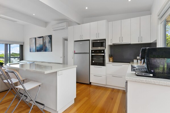 Una cocina moderna con armarios blancos, un refrigerador de acero inoxidable, un horno y un microondas. Hay una barra con taburetes y un ventanal con vistas al exterior.