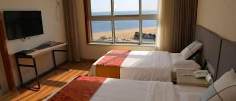 A hotel room with two beds, a desk, a television, and a large window with a beach view.