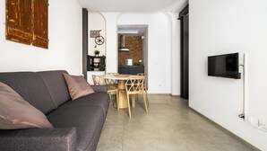 A modern living room with a grey sofa, wooden dining table, and chairs. A TV is mounted on the wall, and a kitchen is visible in the background. - San Michele Apartments - Lapis by Wonderful Italy (Bologna)