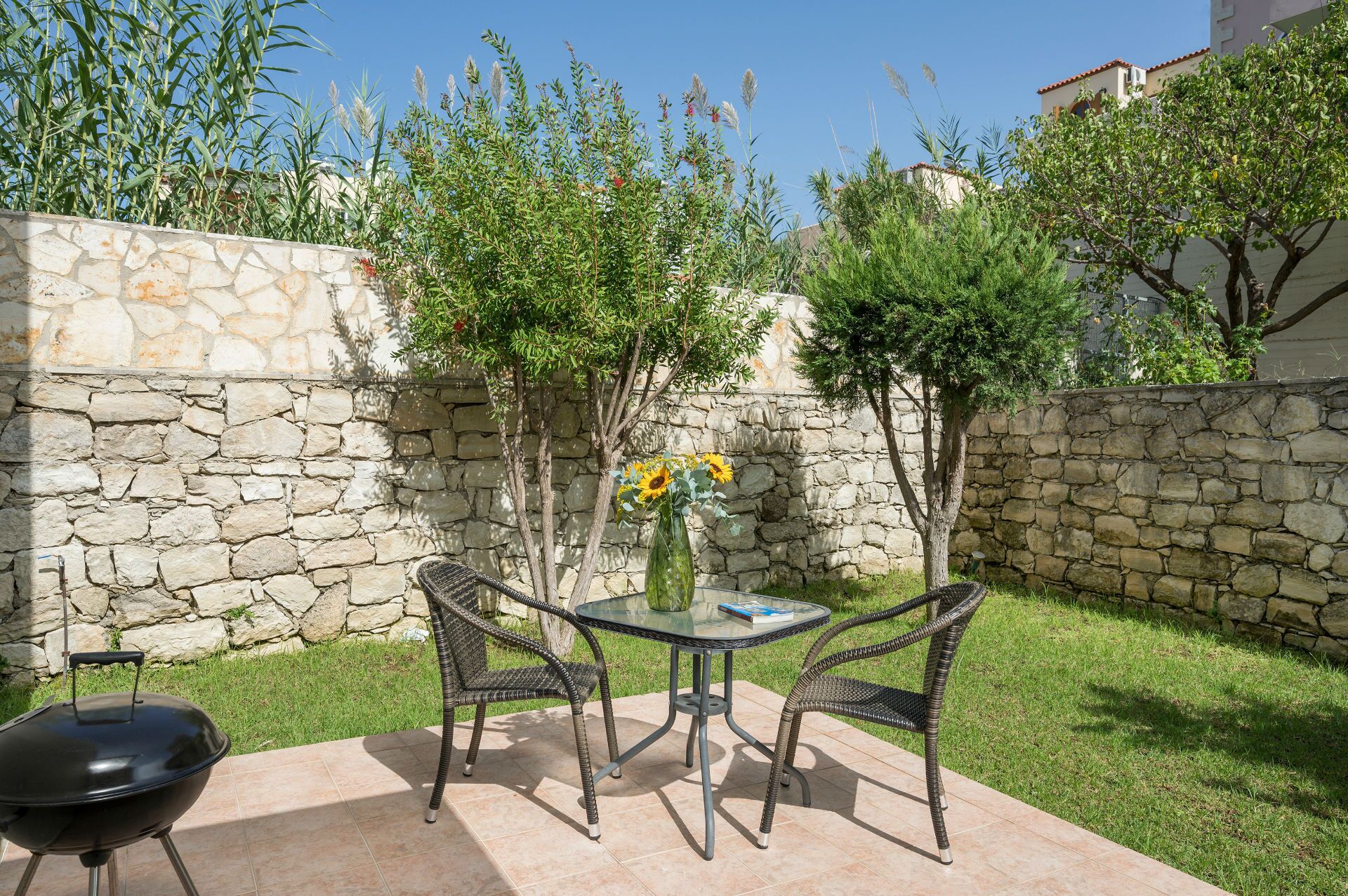 A patio with a round glass table and wicker chairs, a barbecue grill, and a stone wall with greenery.