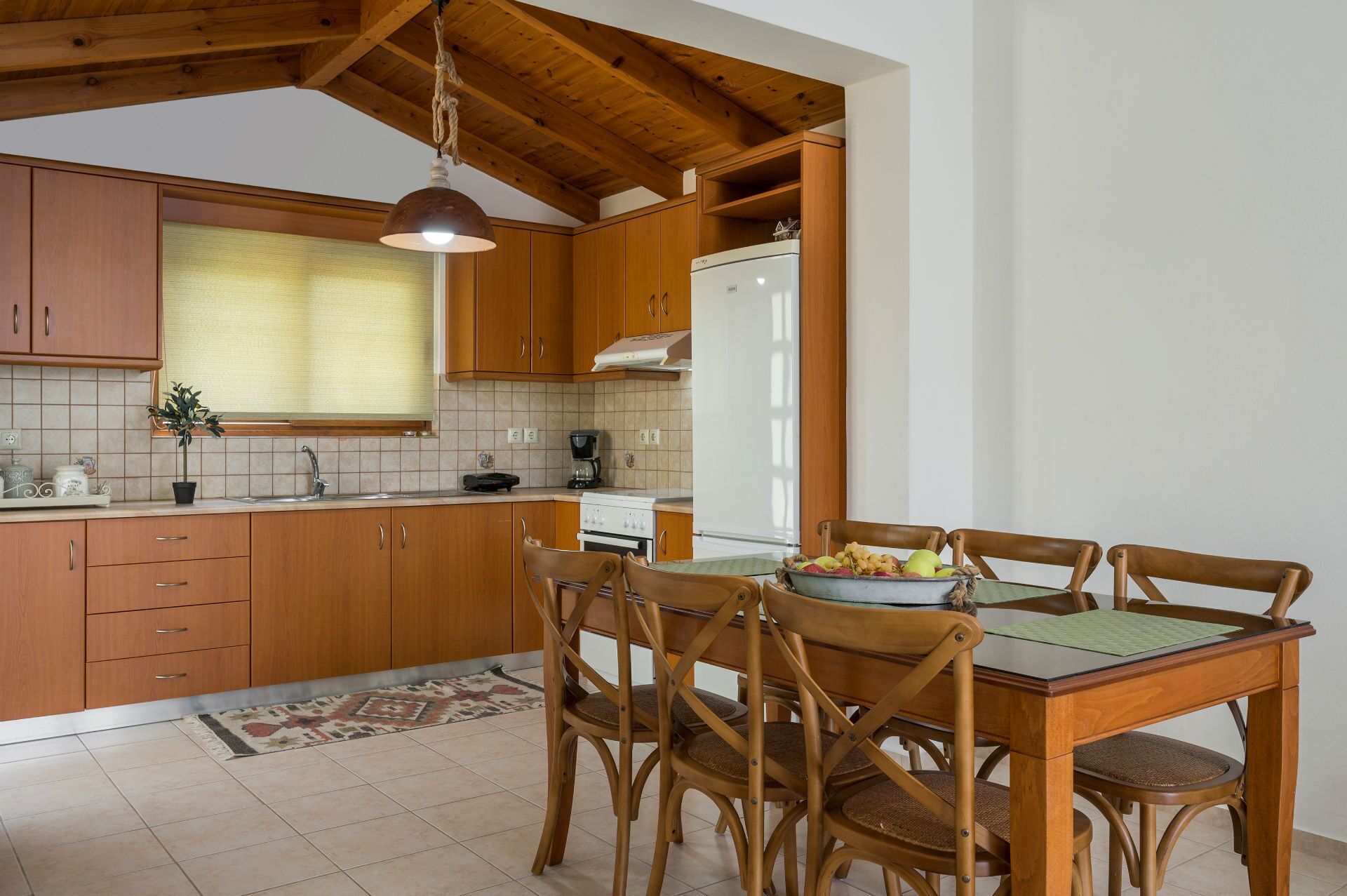 A kitchen with wooden cabinets, a dining table with chairs, a refrigerator, and a hanging light fixture.