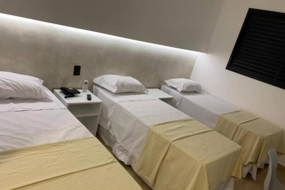 Habitación de hotel con tres camas individuales, cada una con una colcha blanca y una almohada. Hay una mesita con un teléfono y una lámpara entre las camas.