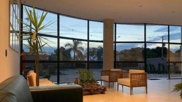 Uma sala de estar ampla com uma grande janela que oferece vista para o exterior. Há um sofá de couro, uma mesa de centro e móveis de vime.